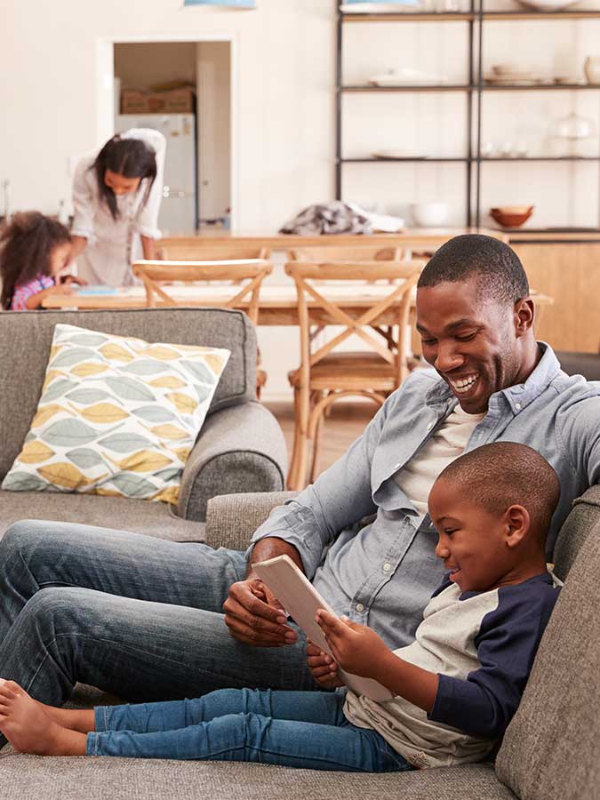 Father and son watching a tablet on the couch, mother and daughter at the table in the background